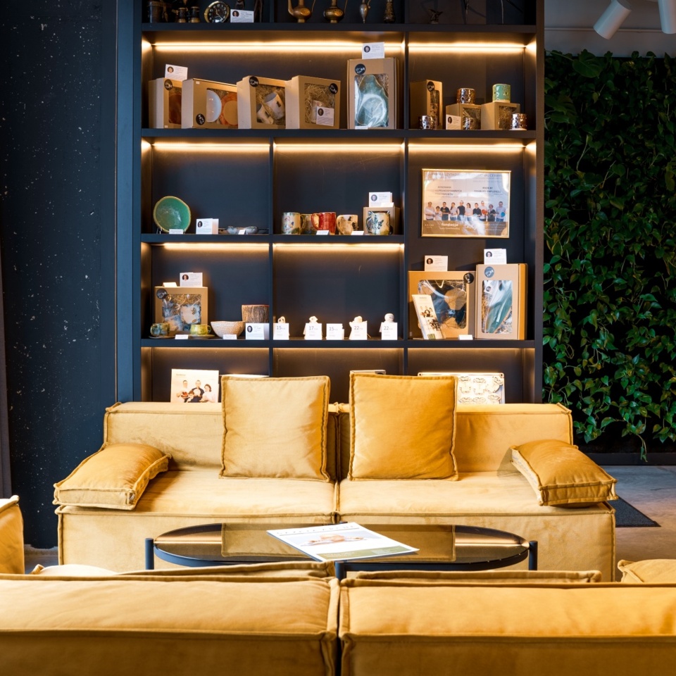 Modern hotel lobby with mustard yellow sofas, backlit shelves displaying local souvenirs, and a lush green plant wall.