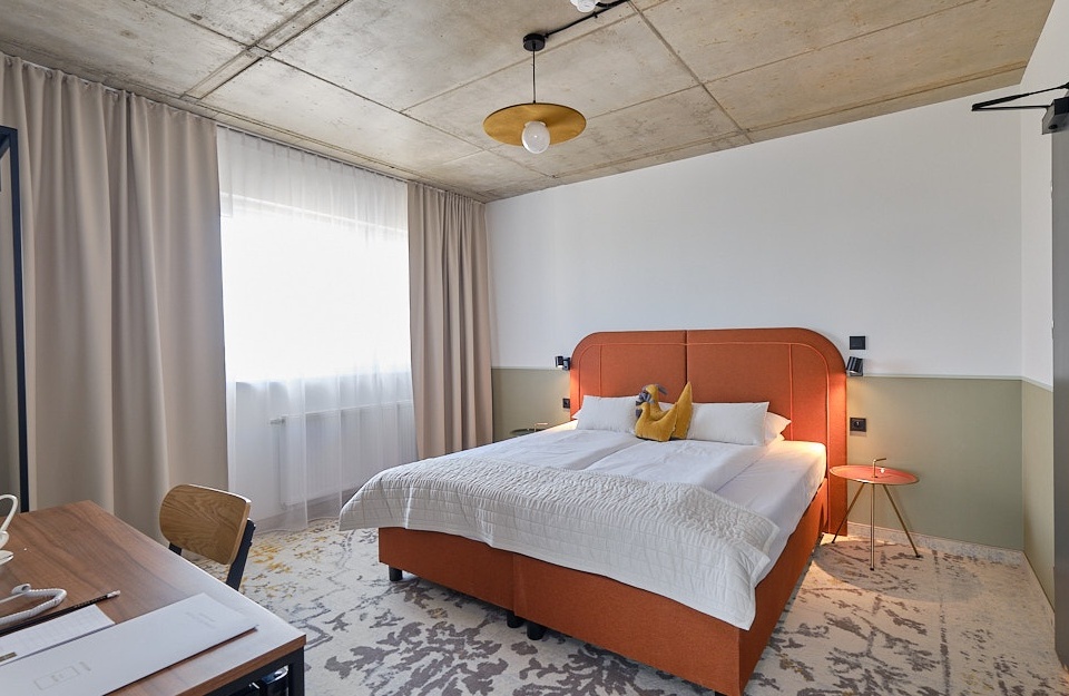 Modern hotel room with large orange double bed, light curtains, carpeted floor, and wooden desk with a lamp