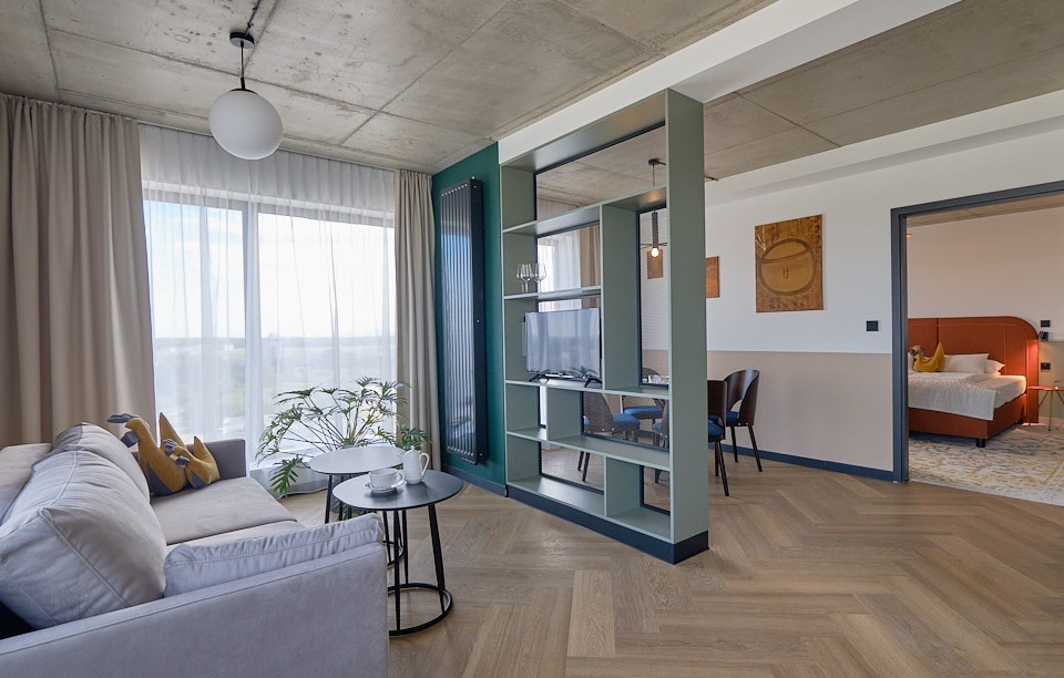 Modern suite with spacious living area, large window, gray sofa, and open bedroom featuring a bed with orange upholstery.