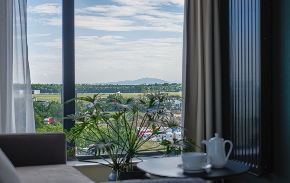 Hotel room with large window overlooking green landscape and distant hills, cozy sofa, and table with coffee set
