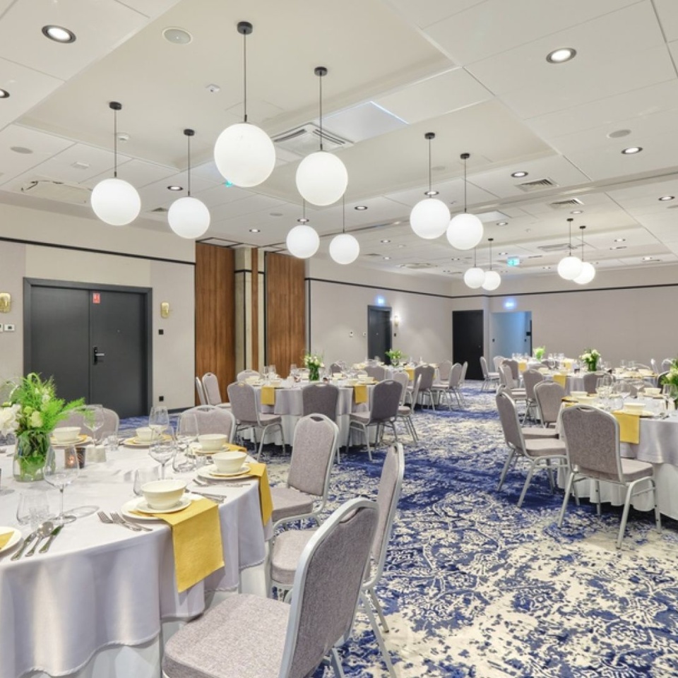 Elegancka sala bankietowa z okrągłymi stołami nakrytymi białymi obrusami i żółtymi serwetkami, oświetlona kulistymi lampami.