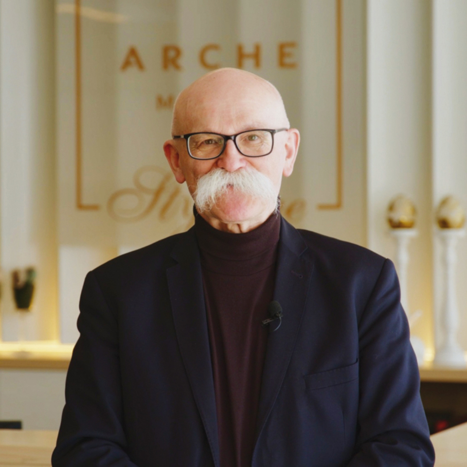 Starszy mężczyzna z okularami i w ciemnym garniturze stoi w eleganckim lobby hotelu z jasnym wnętrzem i dekoracjami