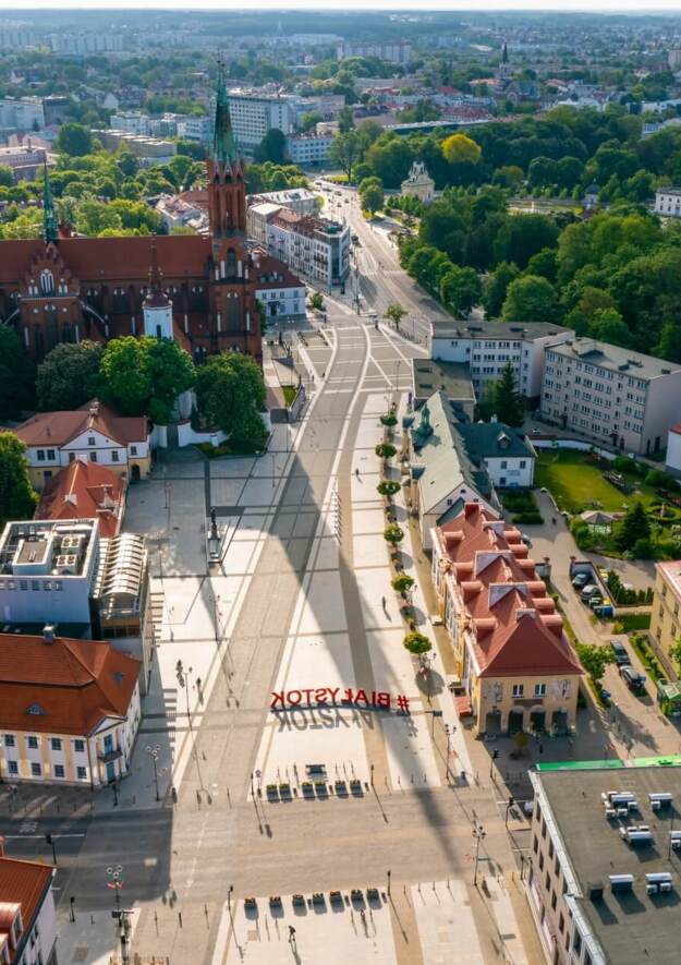 Widok z lotu ptaka na ceglany kościół obok dużego deptaka, zielone parki i hotele z czerwonymi dachami.