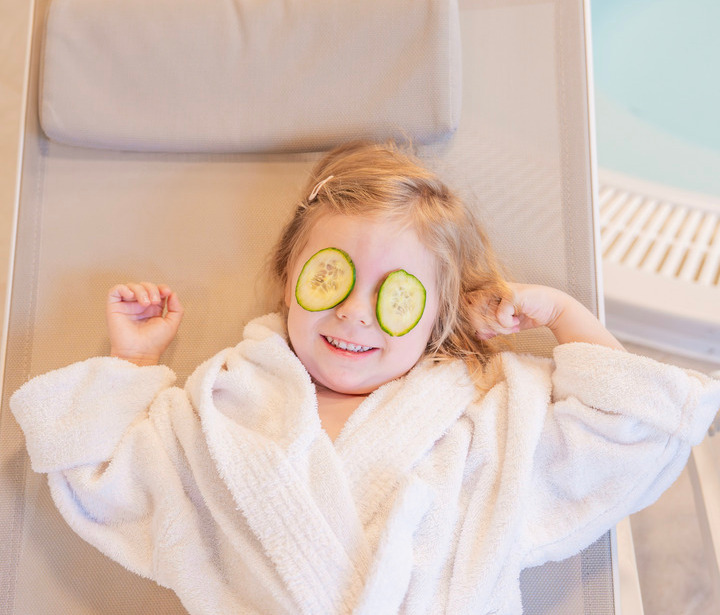 Kind im weißen Spa-Bademantel entspannt auf einer Liege mit Gurkenscheiben auf den Augen neben dem Hotel‑Whirlpool