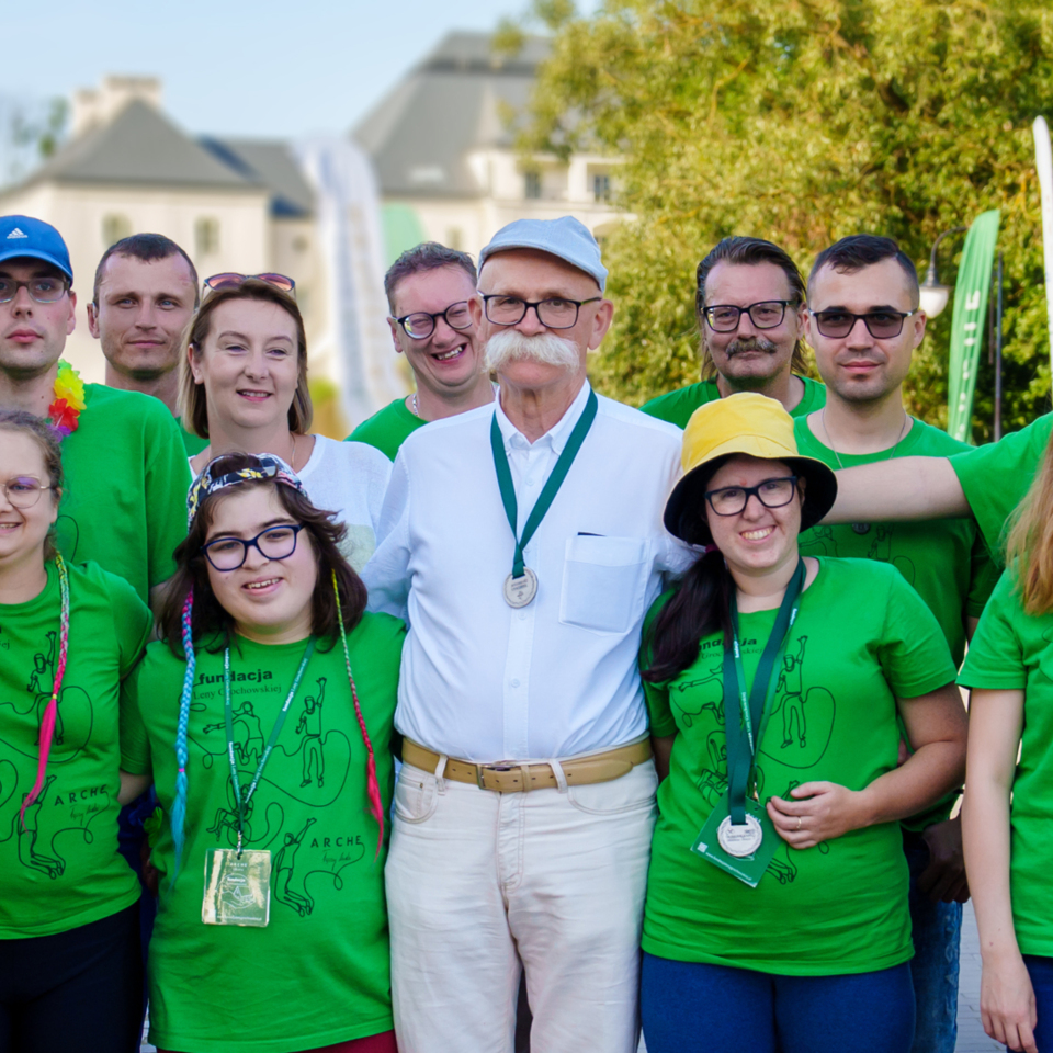 Grupa uśmiechniętych wolontariuszy w zielonych koszulkach na zewnątrz, obok starszy mężczyzna w białej koszuli z medalem, pałac w tle