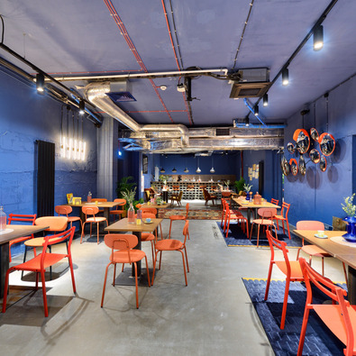 Modern restaurant with blue walls, orange chairs, and industrial ceilings with exposed ductwork and piping.