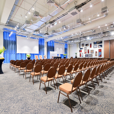 Spacious conference room with rows of brown chairs, projector screen, blue curtains, and modern wall art in bright setting.