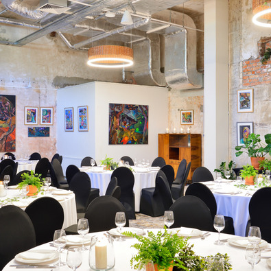 Stylish banquet room with round tables, black chair covers, green plants, and abstract paintings on exposed brick walls.