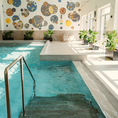 Bright indoor pool with mosaic wall art, potted plants, and sunlit seating area near large windows in a hotel.