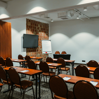 Conference room with rows of wooden tables and brown chairs, flipchart, and wall-mounted TV for presentations.