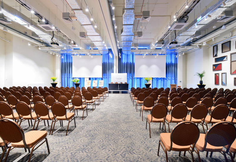 Modern conference room with rows of brown chairs, two projection screens, floral decorations, and a spacious carpeted floor