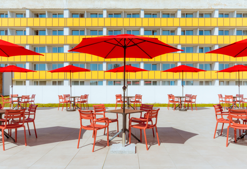 Hotel terrace with red umbrellas and chairs, modern facade featuring yellow panels and balconies.