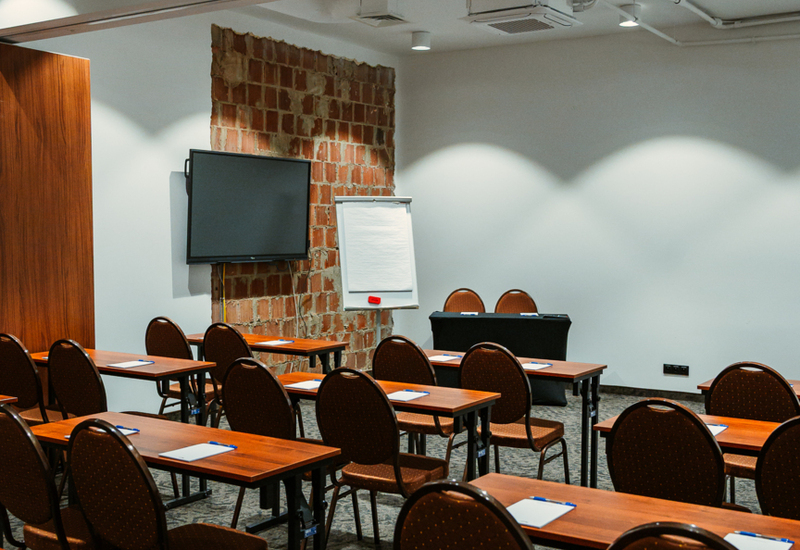 Conference room with rows of wooden tables and brown chairs, flipchart, and wall-mounted TV for presentations.