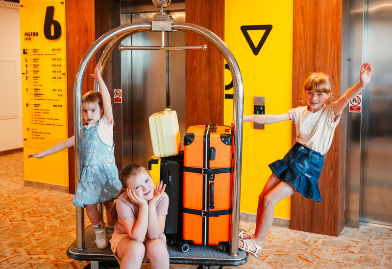 Three children play with a hotel luggage cart carrying bright suitcases in a brightly lit corridor by two elevators.