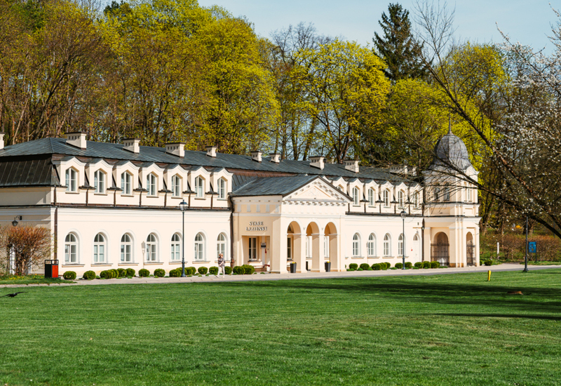 Classic cream-colored hotel building with arches and a tower, surrounded by green lawn and spring trees under clear sky.