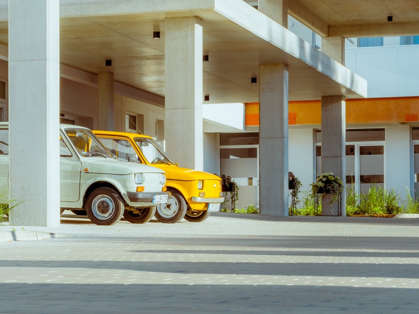 Nowoczesny hotel z kolorowymi balkonami i betonowym zadaszeniem w strefie parkingu gości z dwoma zaparkowanymi samochodami.