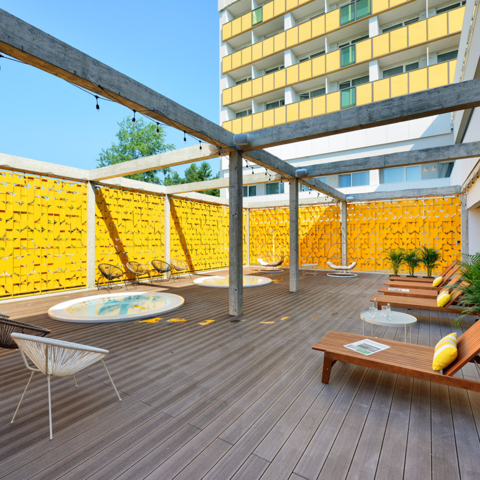 Nowoczesny hotelowy taras z drewnianymi leżakami, jacuzzi, huśtawkami i żółtymi panelami dekoracyjnymi.