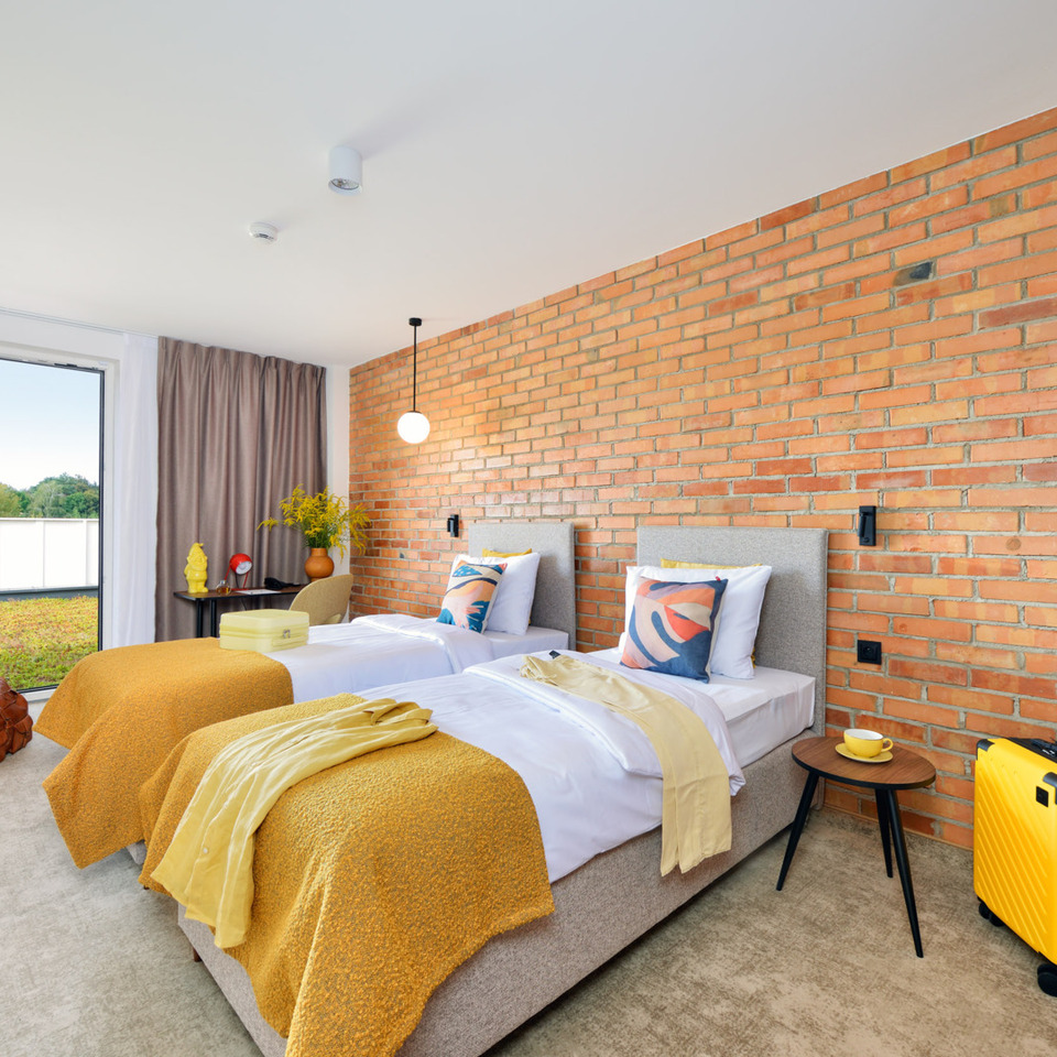 Hotel room with two single beds, exposed brick wall, yellow throws, and a large balcony overlooking greenery.