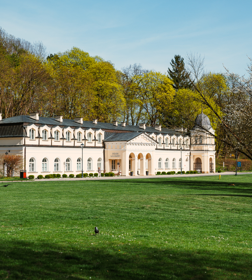 Classic cream-colored hotel building with arches and a tower, surrounded by green lawn and spring trees under clear sky.
