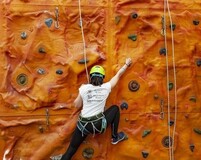 Gość w kasku i uprzęży wspina się po pomarańczowej, krytej ściance wspinaczkowej w hotelu, widoczne liny asekuracyjne.