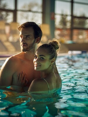 Para relaksuje się w krytym basenie hotelowym z dużymi przeszkleniami, leżakami i ciepłym, relaksującym światłem.