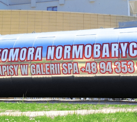 Błyszcząca metalowa komora normobaryczna przed spa, z dużym napisem o zapisach i widocznym numerem telefonu