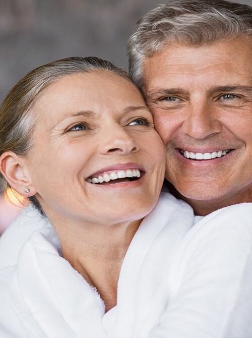 Dojrzała para w białych szlafrokach, uśmiechnięci i przytuleni w strefie relaksu spa oświetlonej świecami