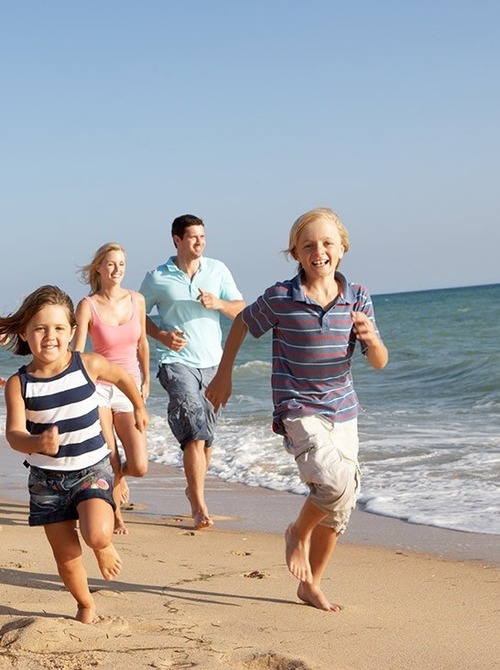 Mehrere Generationen rennen barfuß über sonnigen Sandstrand, lachen am ruhigen blauen Meer.