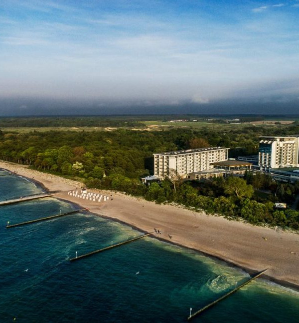 Kompleks hotelowy przy morzu z piaszczystą plażą, drewnianymi falochronami, rzędami białych leżaków i zalesionymi wydmami w tle
