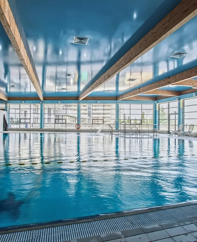 Kryty, ogrzewany basen hotelowy z odkrytymi drewnianymi belkami, błyszczącym niebieskim sufitem, leżakami i dużymi oknami.