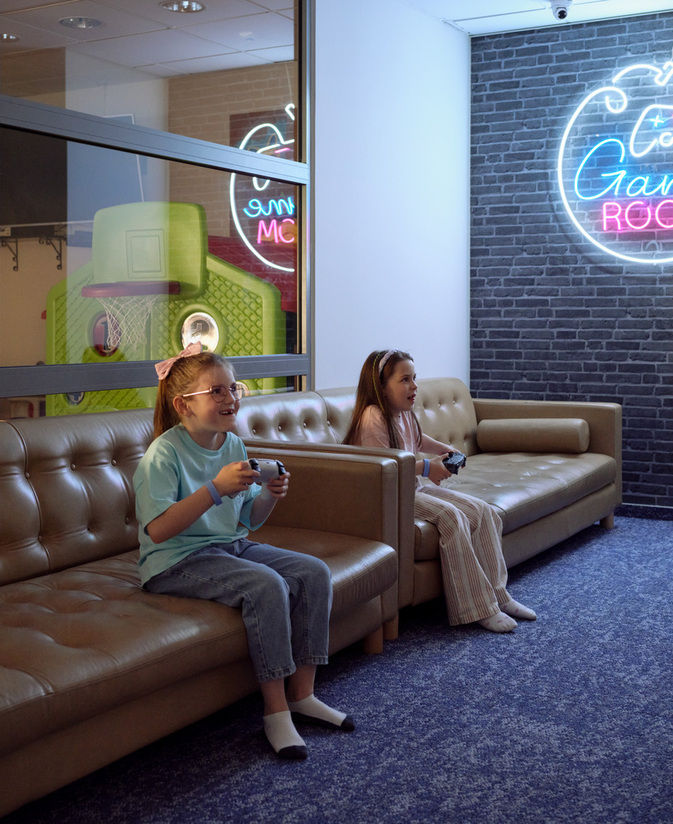 Rodzinny pokój gier z skórzanymi sofami, neonowym napisem i dwiema dziewczynkami grającymi na konsoli.