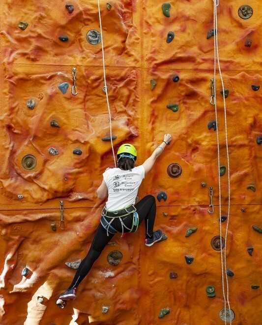 Gość w kasku i uprzęży wspina się po pomarańczowej, krytej ściance wspinaczkowej w hotelu, widoczne liny asekuracyjne.