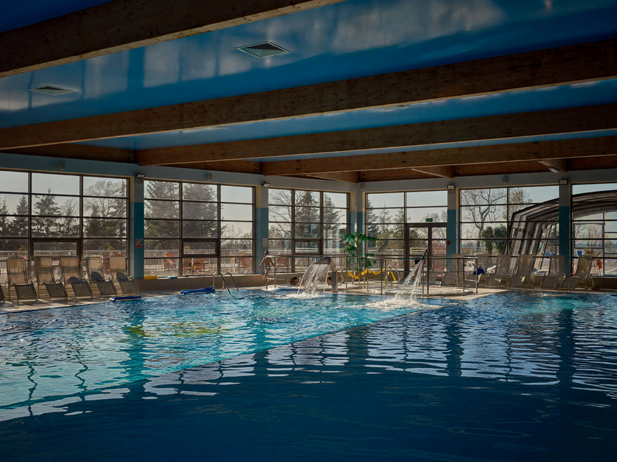 Przestronny kryty basen hotelowy z odsłoniętymi drewnianymi belkami, dużymi oknami, leżakami, kaskadami i gościem wchodzącym do wody.