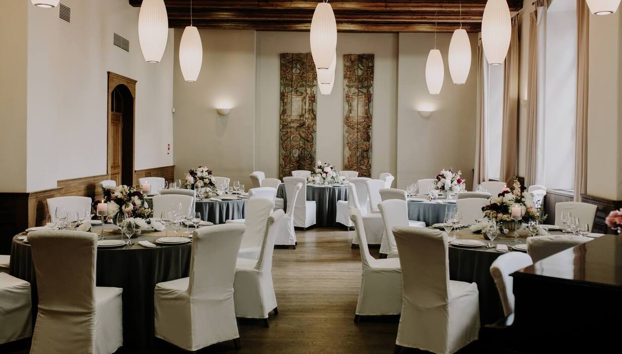 Elegant banquet room with round tables, white chair covers, floral centerpieces, candles, pendant lights and wooden beams.
