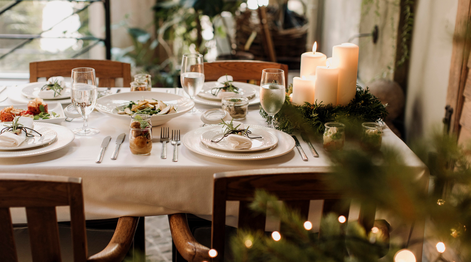 Elegant dining table with white linens, place settings, water glasses, jar appetizers and lit pillar-candle centerpiece.
