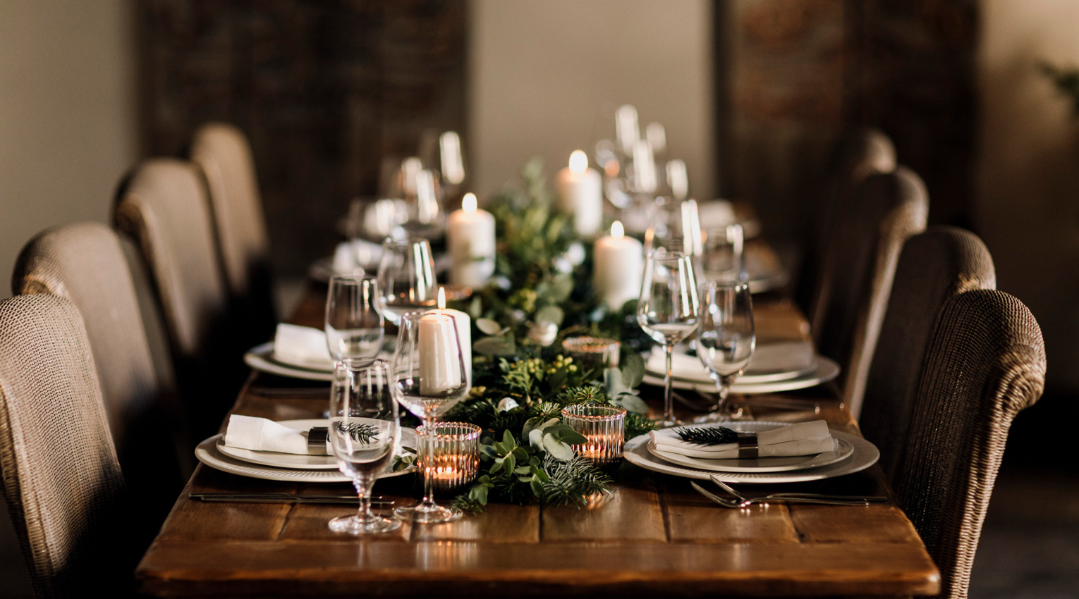 Long wooden table set for an intimate dinner with white plates, wine glasses, lit candles and green foliage centerpiece