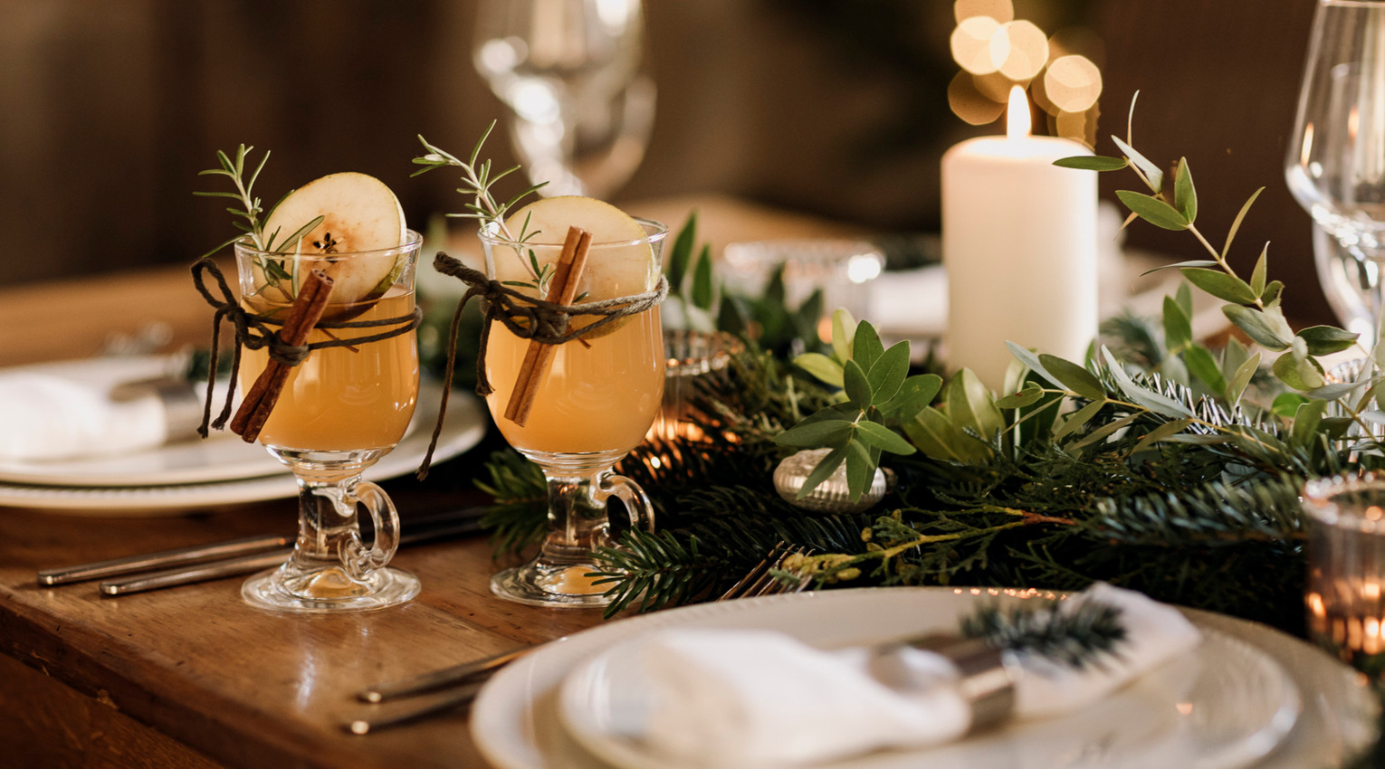 Candlelit holiday table with two warm spiced apple cocktails garnished with cinnamon sticks, rosemary and apple slices.