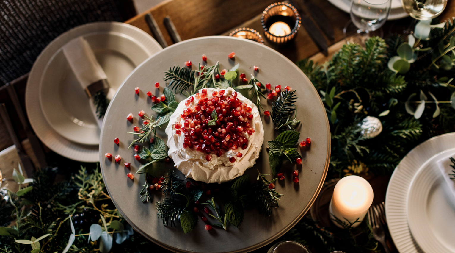Festive pavlova topped with pomegranate seeds on a greenery-decorated platter, candlelit table with plates and wine glasses