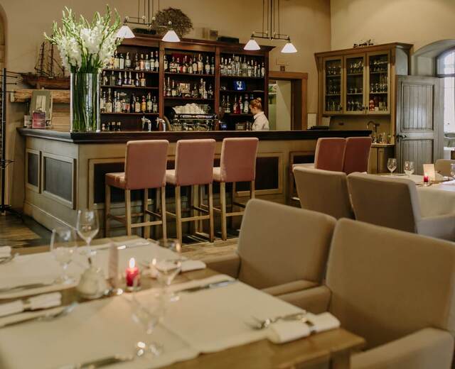 Przytulna hotelowa restauracja z drewnianym barem, wazonem białych kwiatów, różowymi hokerami, nakrytymi stołami ze świecami i barmanem.