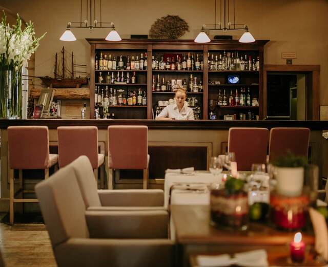 Przytulny bar hotelowy z drewnianym regałem alkoholi, różowymi hokerami, barmanką i świecami na stolikach.