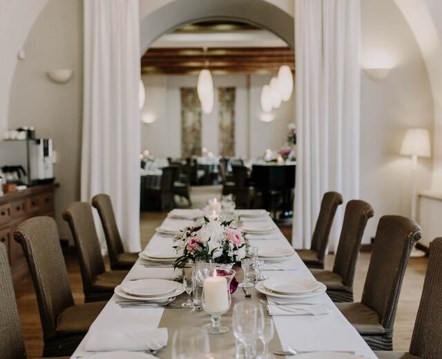 Elegancka sala jadalna z długim stołem w białych obrusach, kwiatowymi aranżacjami, palącymi świecami i obitymi krzesłami pod łukiem.