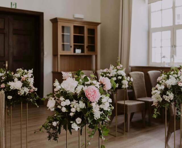 Elegancka sala ceremonii z różowo-białymi kompozycjami kwiatowymi na złotych stojakach, plecionymi krzesłami i światłem z dużego okna.
