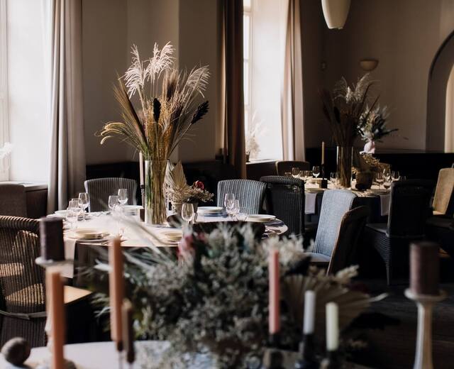 Przytulna jadalnia hotelowa z okrągłymi stołami, wiklinowymi fotelami, suszonymi kompozycjami i eleganckim nakryciem.
