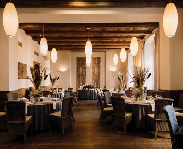 Elegancka sala restauracyjna z okrągłymi stołami w szarych obrusach, wiklinowymi krzesłami, wiszącymi lampami, drewnianym stropem i świecami.