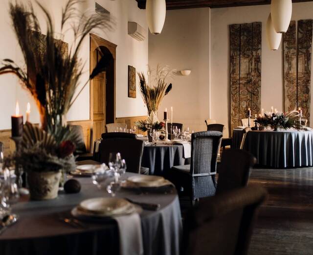 Elegancka sala jadalna z okrągłymi stołami w szarych obrusach, świecami, kwiatowymi dekoracjami, plecionymi krzesłami i drewnianą boazerią
