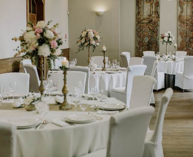 Sala bankietowa z okrągłymi stołami, białymi obrusami i pokrowcami, wysokimi bukietami, świecami i eleganckim nakryciem.