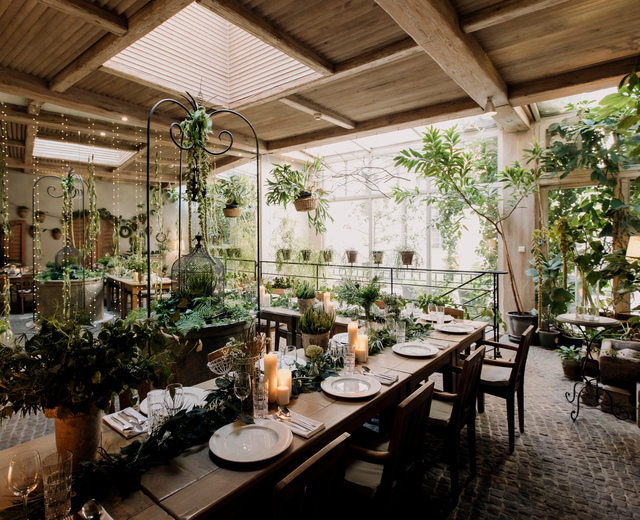 Sala restauracyjna w oranżerii pełna roślin, z drewnianymi belkami, świetlikami, wiszącymi koszami i stolikami przy świecach.