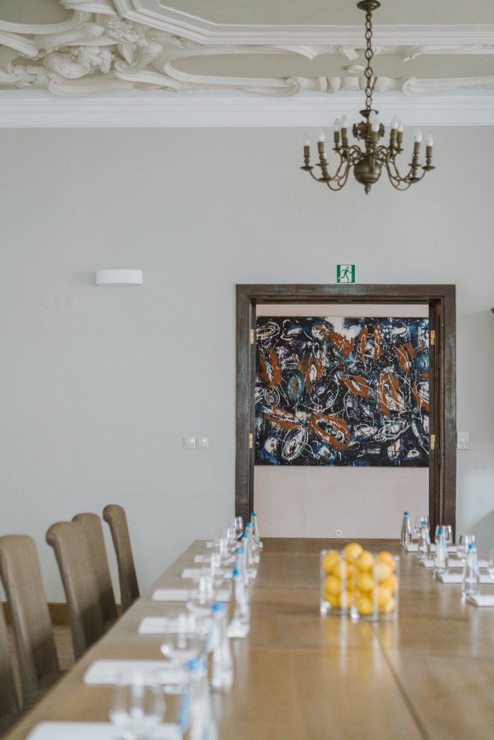 Elegancka sala konferencyjna z długim drewnianym stołem przygotowanym z butelkami wody, szklankami i notatnikami, żyrandolem i misą cytryn