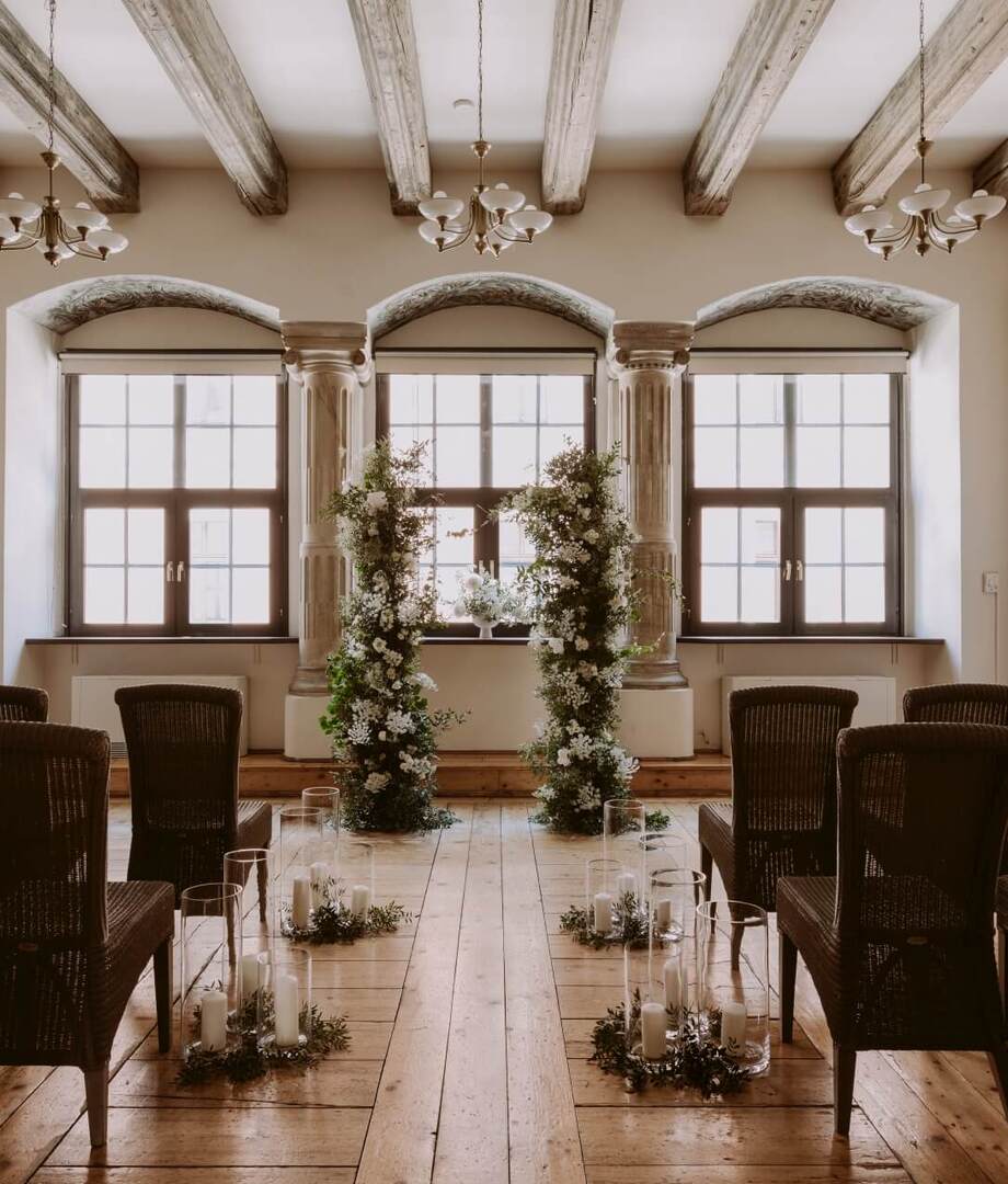 Intimate ceremony room with wooden beams and floor, arched windows, floral arch, candlelit aisle and wicker guest chairs.