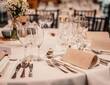 Elegant banquet table with white linens, multiple wine glasses, folded napkins, silver cutlery and small daisy centerpiece.